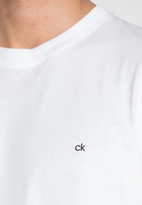 Ingrandimento del colletto di una maglietta bianca semplice con un piccolo logo "ck" nero sul lato sinistro. Texture del tessuto morbida visibile.