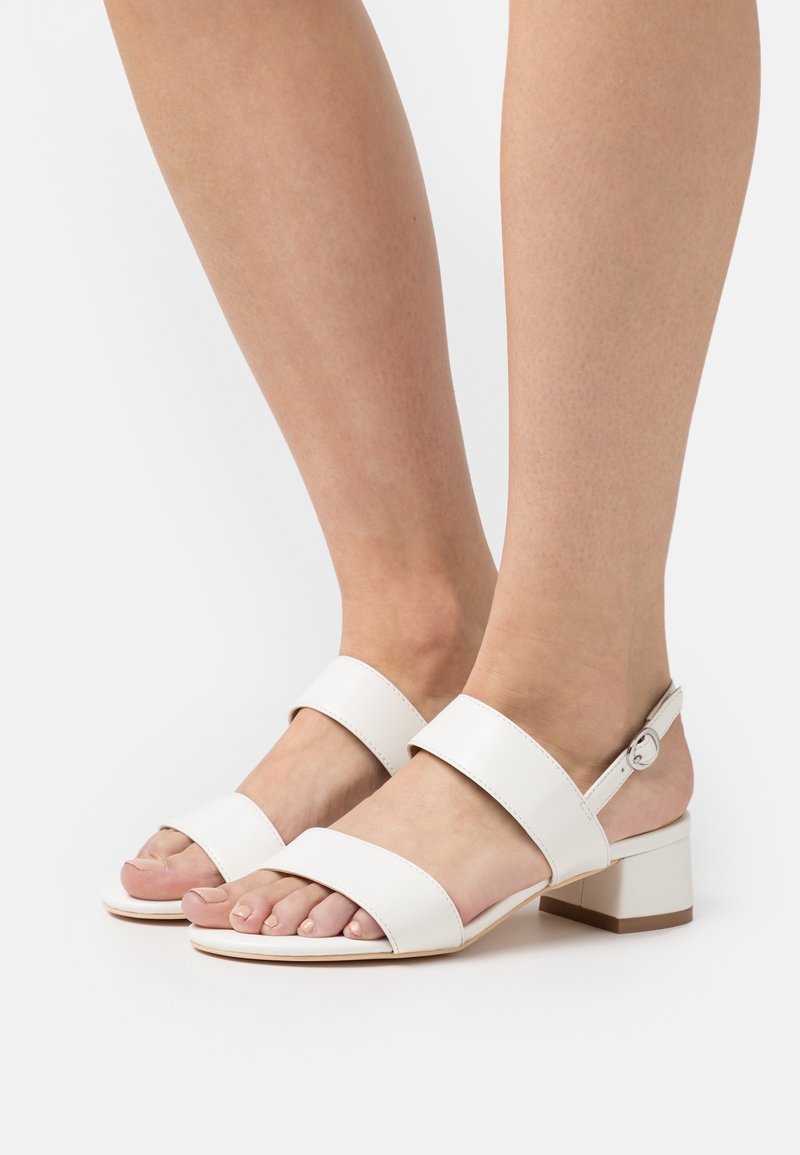 Pieds portant des sandales blanches à petit talon avec deux larges brides et une petite bride à boucle autour de la cheville sur un fond uni.
