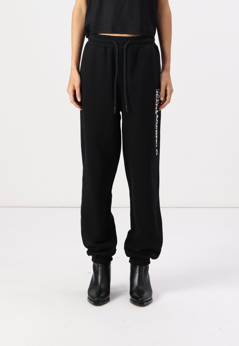 Pantalon de survêtement noir avec taille élastique et cordon de serrage. Marques latérales en blanc. Jambe fuselée avec poignets resserrés et texture douce.