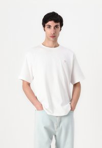 Camiseta blanca de manga corta con cuello redondo, fabricada en suave algodón, presenta un pequeño emblema color óxido en el pecho. Jeans de color azul claro.