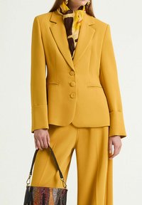 Blazer jaune sur mesure à trois boutons, col affûté et manches structurées. Assorti à un pantalon assorti et une écharpe à motifs. Sac à main noir.