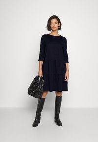 Tiefblaues Baumwollkleid mit Dreiviertelärmeln und gestuften Design. Kombiniert mit einer strukturierten schwarzen Handtasche und knielangen schwarzen Stiefeletten mit Absatz.