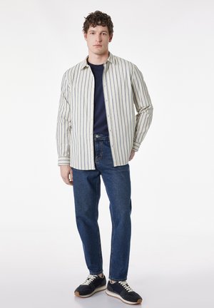 Jonge man met donkere sneakers, blauwe jeans, marineblauw t-shirt en een open wit overhemd met blauwe verticale strepen, staand tegen een effen achtergrond.
