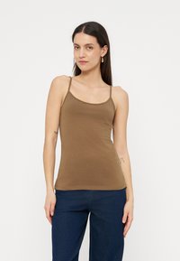 Γυναίκα που φοράει καφέ τοπάκι με λεπτές τιράντες και σκούρο μπλε παντελόνι, στέκεται μπροστά σε λευκό φόντο.