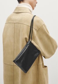 Sort læder crossbody taske med slank design, glat tekstur og to lange stropper, båret over en beige ruskind frakke.