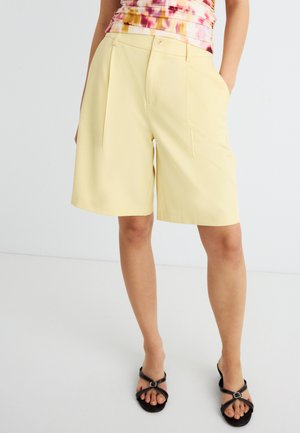 Nő világossárga, térdig érő, szabott rövidnadrágot visel oldalzsebekkel és fekete pántos szandált, fehér padlón áll.