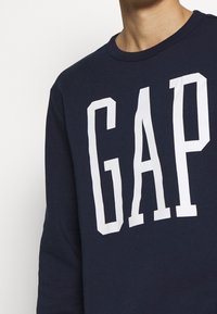 Felpa navy con colletto tondo. Presenta un grande logo bianco "GAP" sulla parte anteriore. Realizzata in un morbido tessuto testurizzato.