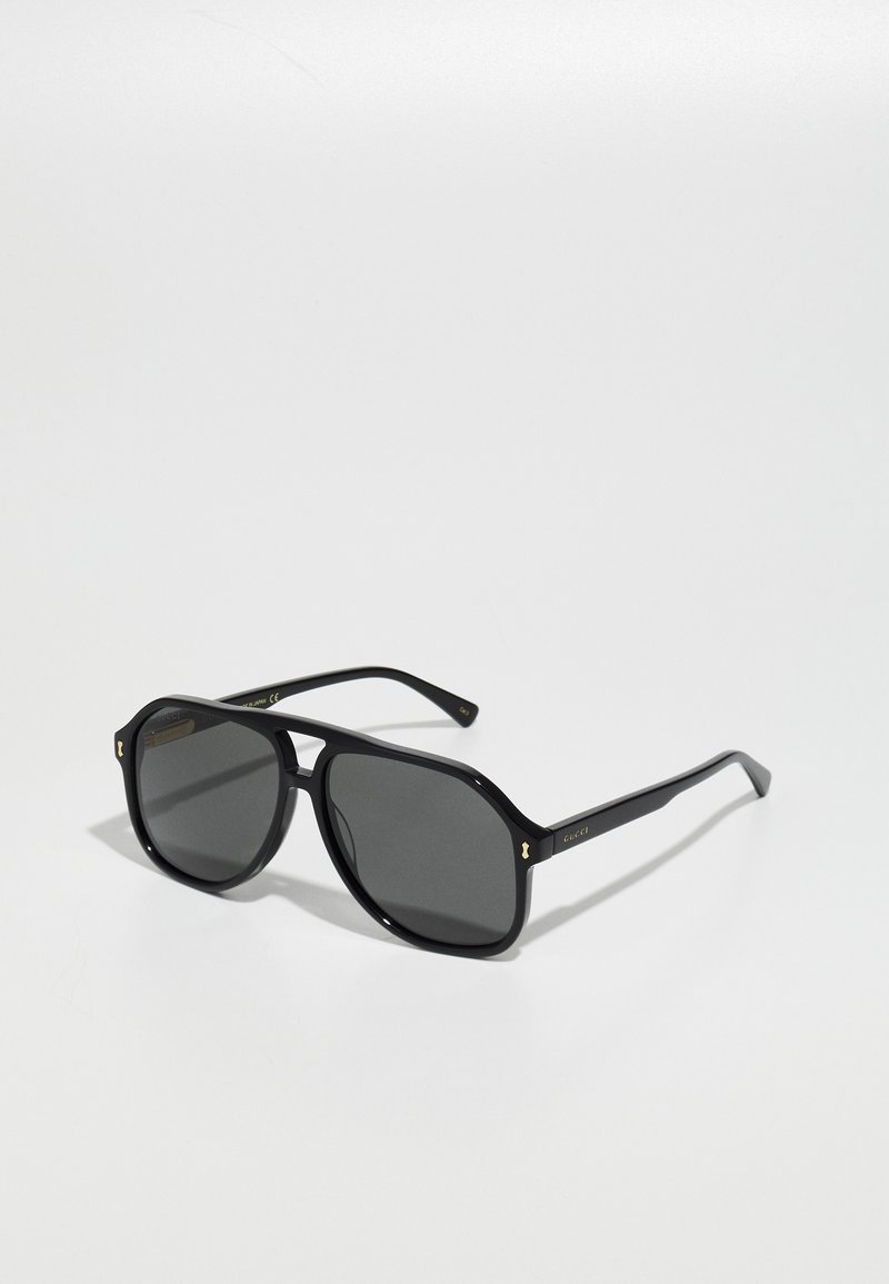 Gafas de sol negras con montura octogonal, lentes oscuros y acentos dorados en las patillas, hechas de un material plástico brillante.
