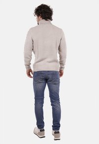 Maglione beige a collo alto lavorato a maglia con un motivo strutturato, abbinato a jeans in denim e sneakers bianche. Visto da dietro.