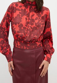 Rød bluse med blomstermønster i forskellige nuancer af rød, høj afslutning ved halsen og lange ærmer, stylet med en bordeaux læderskørt.