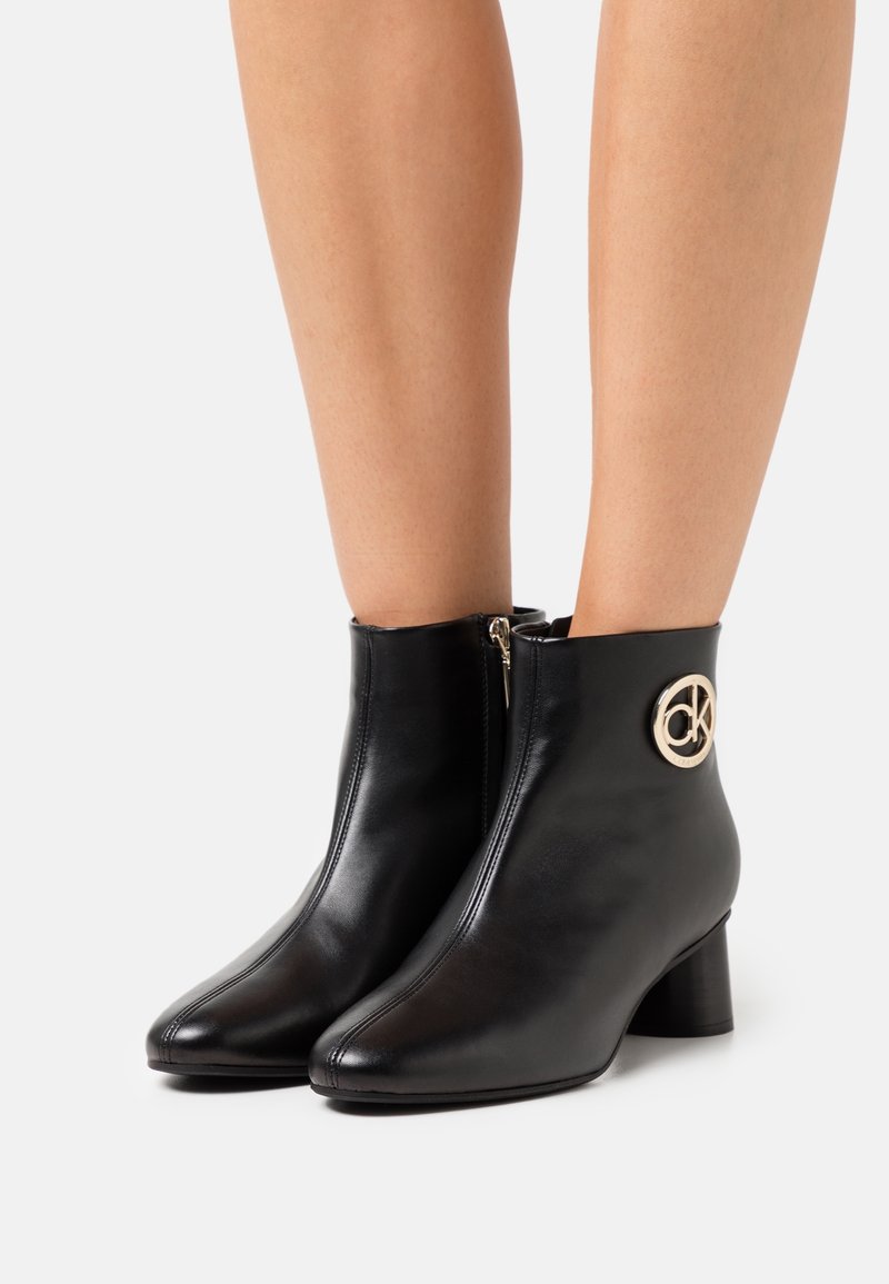 Stivali ankle in pelle nera con chiusura a zip, punta tonda e tacco basso a blocco, con un accento del logo circolare dorato.