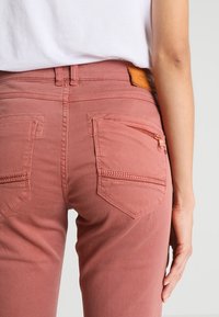 Džínsy v barvě starorůžové s rovnym střihem, obsahující dvě zadní náprsní kapsy a kapsu na zip. Hladká textura s koženou nášivkou.