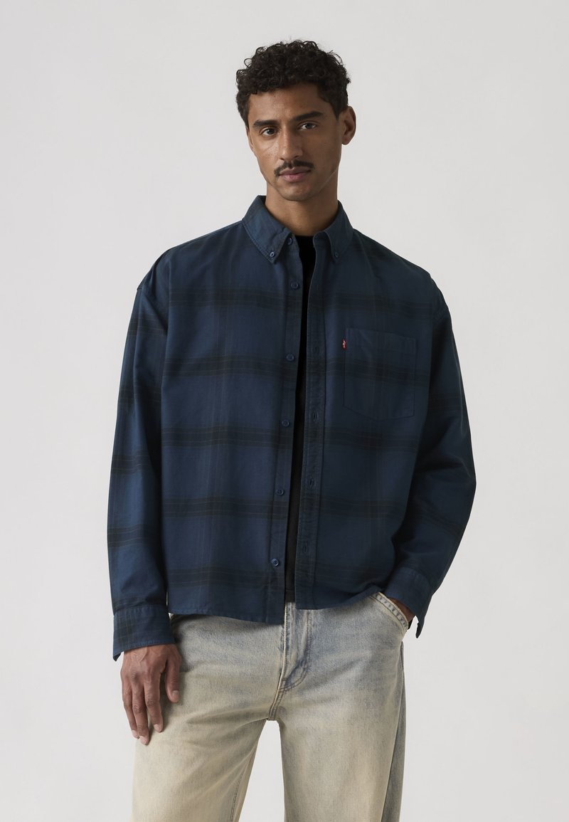 Muškarac odjeven u tamnoplavu košulju s gumbima preko crnog topa i svijetlih traperica, stoji s jednom rukom u džepu nasuprot jednobojnoj pozadini.