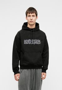 Melnas kokvilnas kapuce ar priekšējo kangaru kabatu un loģotipa tekstu novecojušā stilā. Apvienota ar gaiši pelēkiem sporta biksēm.