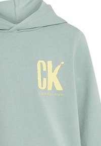 Felpa blu chiaro realizzata in tessuto morbido, con logo giallo "CK" e testo "Calvin Klein Jeans" in un carattere pulito sul davanti.