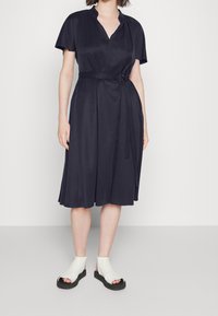 Mujer vestida con un vestido negro de longitud hasta la rodilla, con cinturón y mangas cortas, y sandalias blancas de punta abierta, sobre un fondo blanco liso.