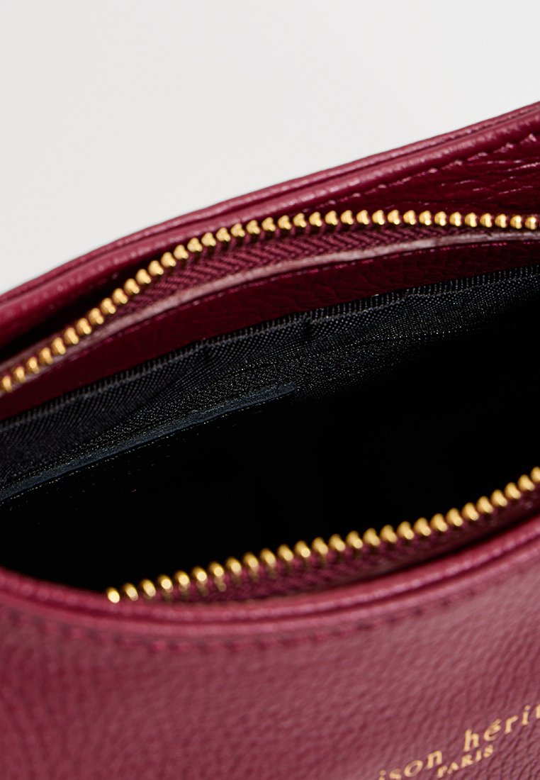 Pochette en cuir bordeaux avec une surface texturée, fermeture éclair dorée et doublure intérieure noire. Présente un marquage minimal à l'extérieur.