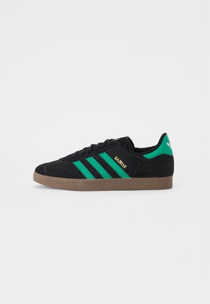 Ténis pretos de camurça com três riscas verdes, uma sola de borracha castanha e a marcação em dourado "Gazelle" na lateral. Design clássico de cano baixo.