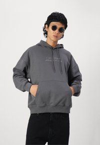 Grijze pullover hoodie met een voorzak, trekkoord in de capuchon en witte tekst op de borst. Het heeft een zachte, gladde textuur. G gedragen met zwarte denim.