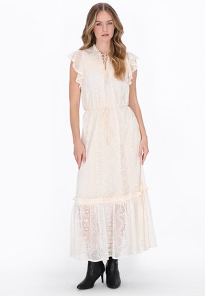 Kobieta w długiej, kremowej koronkowej sukience z falbankowymi rękawami i czarnych, wysokich botkach, stojąca na białym tle.