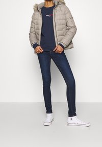 Osoba oblečená v béžové zimní bundě s kapucí, námořnické mikině Tommy Jeans, tmavých slim jeanech a bílých teniskách Converse s vysokým střihém.