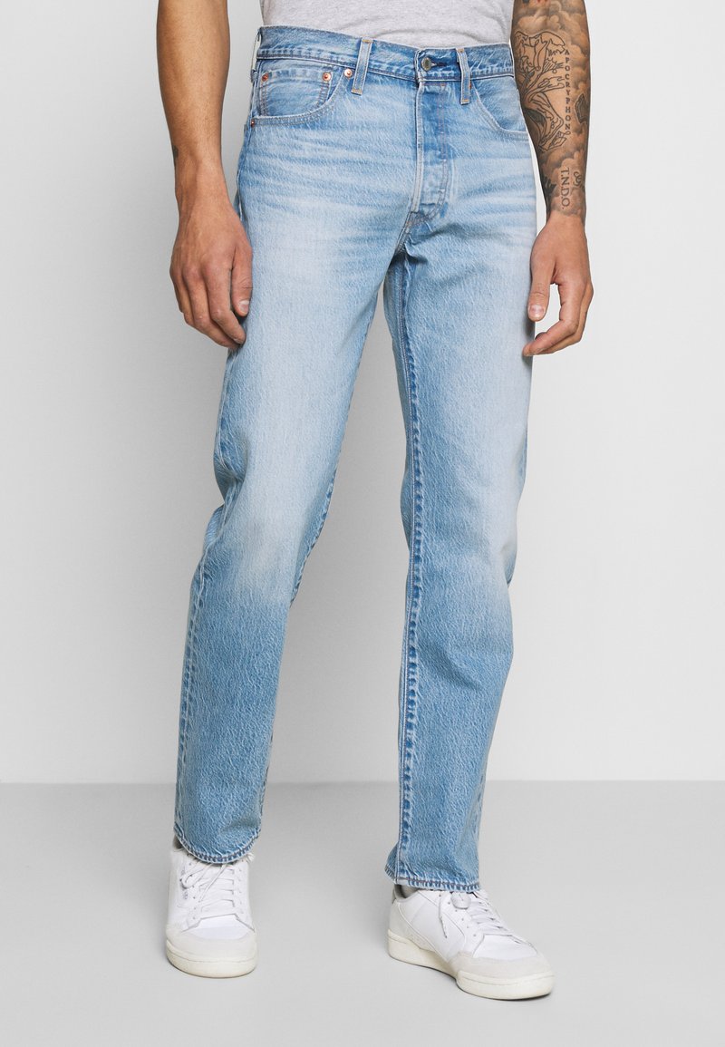 Jeans de mezclilla azul claro con corte recto, cinco bolsillos y herrajes metálicos. Combinados con zapatillas blancas. Tela suave y texturizada.