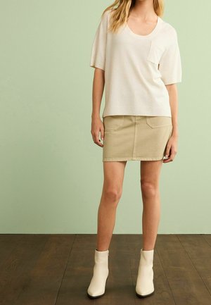 Kobieta ubrana w kremowy top z krótkim rękawem, beżową mini spódnicę i białe botki, stojąca na drewnianej podłodze na tle jasnozielonej ściany.