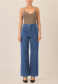 Brauner Body mit dünnen Trägern, kombiniert mit hochgeschnittenen, weit geschnittenen blauen Jeans. Schwarze spitze Absätze betonen das Outfit.