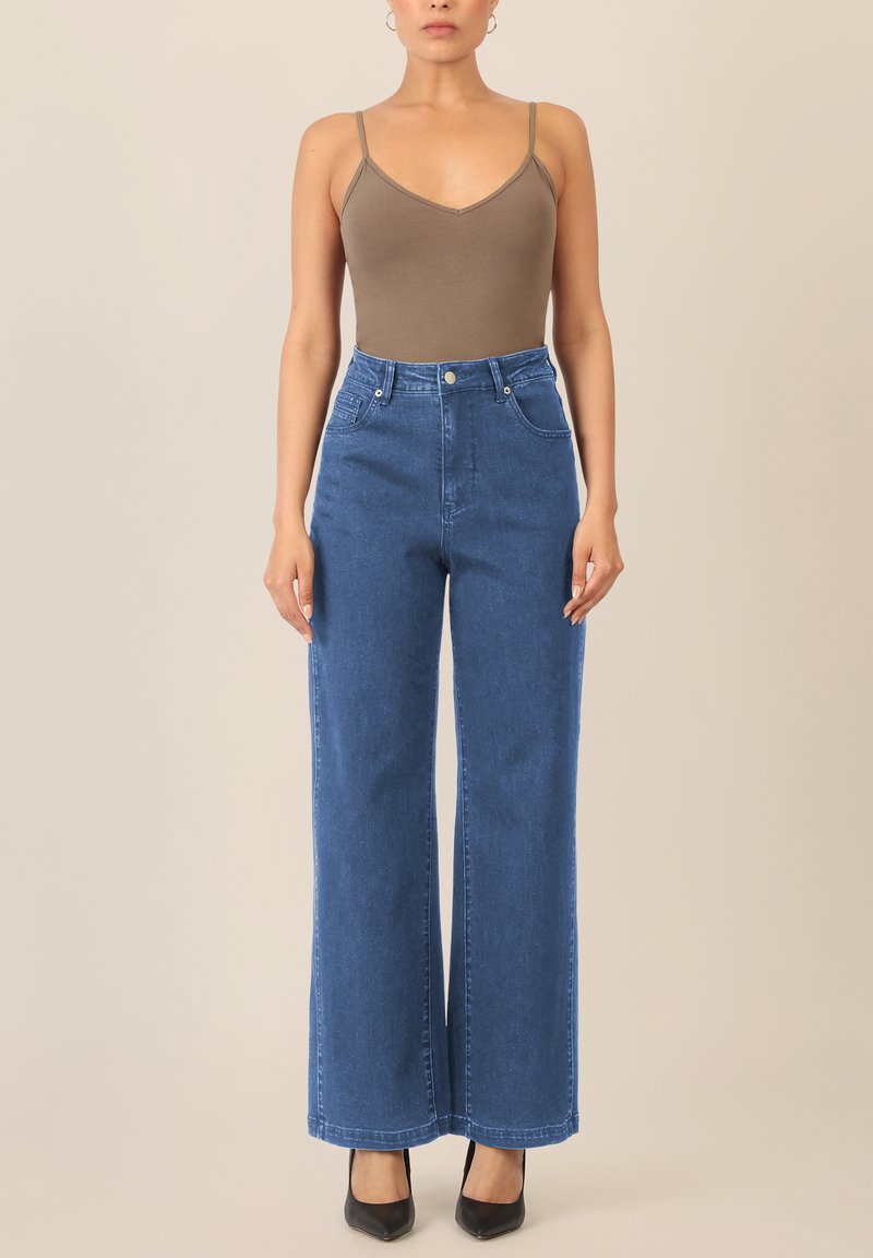 Brauner Body mit dünnen Trägern, kombiniert mit hochgeschnittenen, weit geschnittenen blauen Jeans. Schwarze spitze Absätze betonen das Outfit.