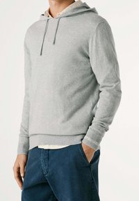 Felpa con cappuccio grigio chiaro realizzata in tessuto morbido, con coulisse, polsini e orlo a coste, indossata sopra una camicia chiara con colletto.