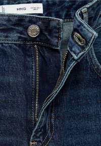 Jeans de mezclilla azul oscuro con cierre de cremallera y botón de metal. Las costuras son visibles a lo largo de las costuras. La etiqueta de cuidados está adjunta en la cinturilla.