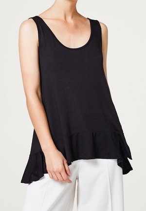 Femme portant un haut noir sans manches ample avec un ourlet à volants et un pantalon blanc à jambes larges, debout devant un fond clair uni.