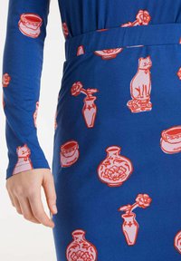 Vestido de tela azul con un estampado de gatos rojos, jarrones y flores. Mangas largas y diseño ceñido en la cintura. Textura suave.
