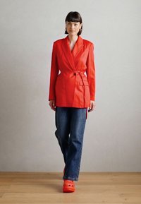 Rdeč elegantni blazer z zaponko v pasu, v kombinaciji z ohlapnimi modrimi džinsami in oranžnimi klobuki. Gladka tkanina in strukturirana silhueta.
