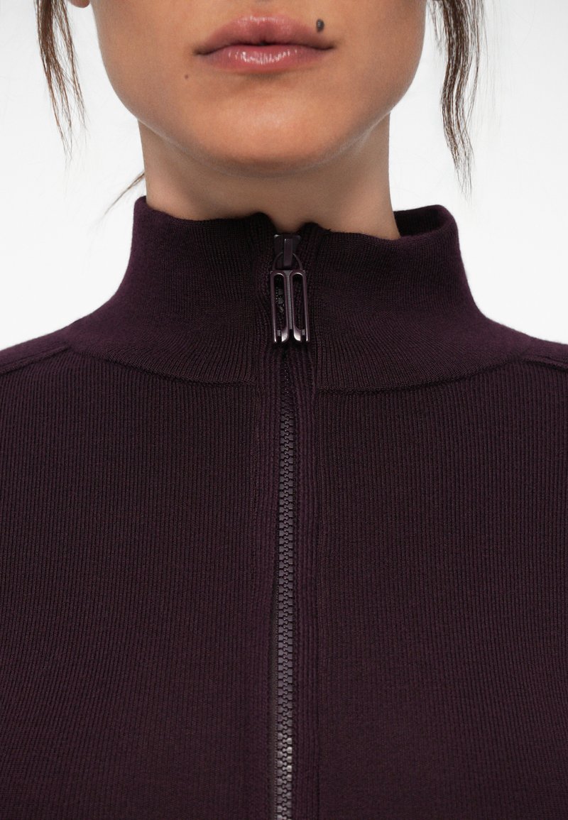 Lila zip-up tröja med ribbad textur och hög krage, med en central dragkedja med svarta detaljer. Inga ytterligare detaljer synliga.