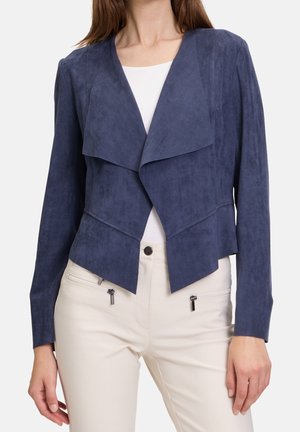 Kobieta ubrana w granatową, otwartą kurtkę z drapowanymi klapami i kremowe spodnie z kieszeniami na zamek.