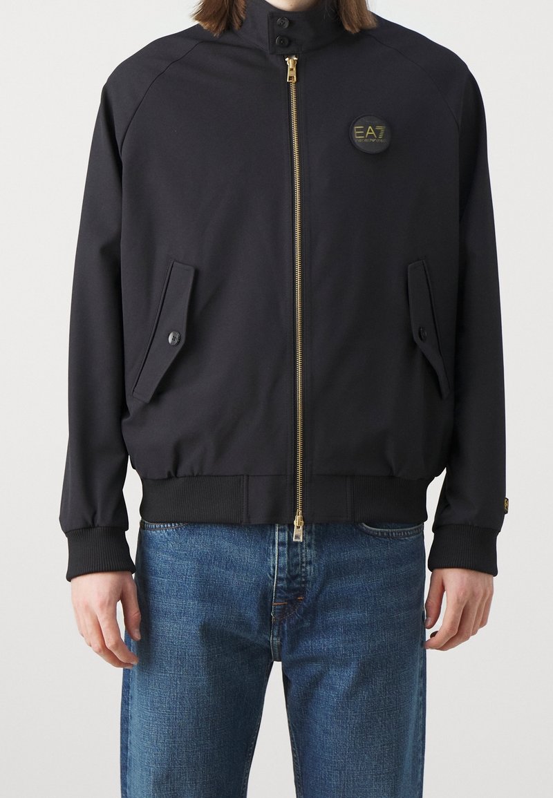 Homme portant une veste noire zippée avec des poches à rabat boutonnées et un petit logo EA7 sur la poitrine gauche, associée à un jean bleu.