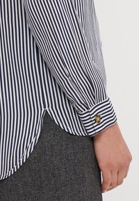 Blusa a righe blu navy e bianche con orlo arrotondato, dotata di polsini con bottone e una texture di tessuto liscia. Indossata con un indumento grigio testurizzato.