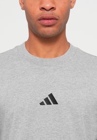 Detailný záber muža v svetlo sivej tričku s kulatým výstrihom, na ktorom je uprostred hrude malé čierne geometrické logo.