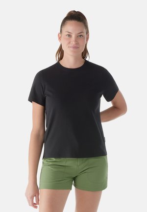 Femme aux cheveux bruns attachés, portant un t-shirt noir uni à manches courtes et un short vert, debout avec une main derrière le dos.