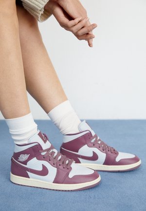 Nogi w białych skarpetkach i fioletowo-białych butach Nike Air Jordan na niebieskim dywanie, ręce splecione nad kolanami.