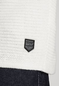 White knit sweater with a textured pattern and a black leather logo patch. Paired with dark denim jeans displaying white stitching.