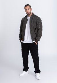 Kurtka bomber w kolorze oliwkowej zieleni z zamkiem błyskawicznym, ściągaczami na mankietach i bocznymi kieszeniami, noszona na białej koszulce i czarnych spodniach bojówkach oraz białych sportowych butach.