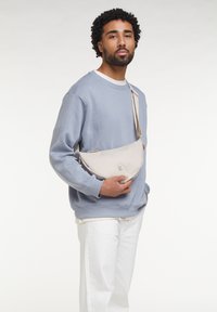 Sac bandoulière beige en tissu doux, avec une forme arrondie et un logo discret. Porté sur l'épaule avec un sweat-shirt bleu décontracté.