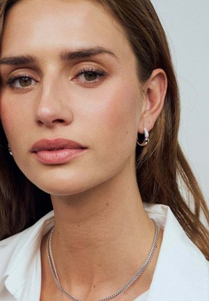 Sølvøreringe med hoops og lagdelte sølvhalskæder supplerer en lysefarvet skjorte, som rammer et neutralt makeup-look og en glat hudtekstur.