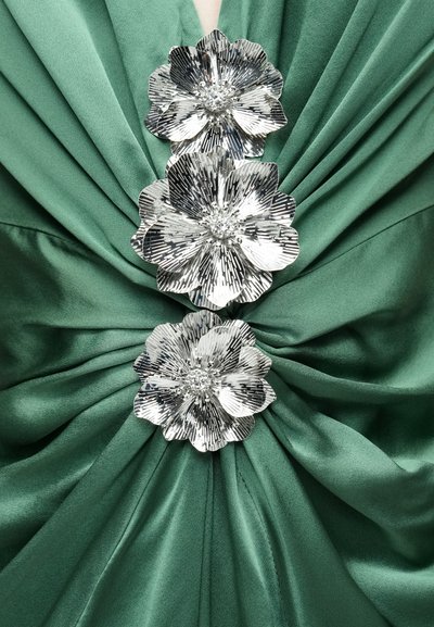 Material satin verde cu pliuri drapate, decorat cu trei ornamente florale argintii, cu petale texturate și un accent central de cristal.