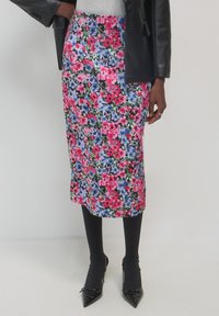 Personne portant une jupe midi à fleurs avec des fleurs roses, bleues et vertes, des collants noirs, des escarpins noirs à bout pointu, un haut gris et une veste en cuir noire.
