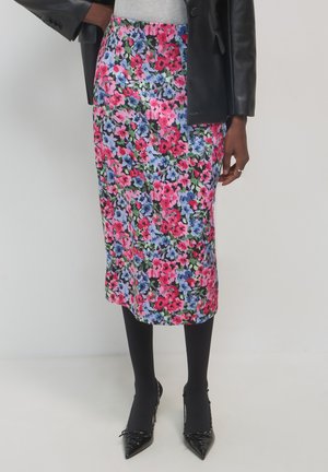 Personne portant une jupe midi à fleurs avec des fleurs roses, bleues et vertes, des collants noirs, des escarpins noirs à bout pointu, un haut gris et une veste en cuir noire.
