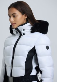 Gepolsterte Pufferjacke in Weiß und Schwarz, mit hohem Kragen, Kunstfellbesatz, Reißverschluss vorne und einem strukturierten schwarzen Gürtel.