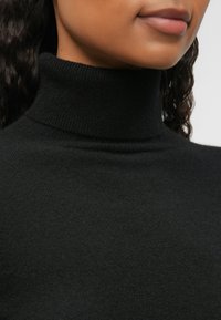Fekete garbó pulóver, lágy, textúrált anyagból, magas nyakkal és sima felülettel, amely csak a nyakat és a felső mellkast ragadja meg.